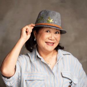 Woman with a grey blouse holding her hat with her right hand against a dark grey background.