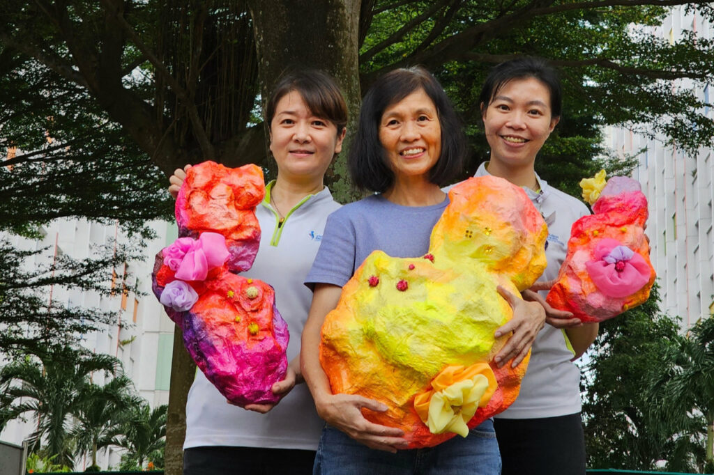 Senior collaborator, Agnes Tan standing in the middle with 2 staff from Yishun Health on her left and right, holding colourful paper mache artwork in their hands.