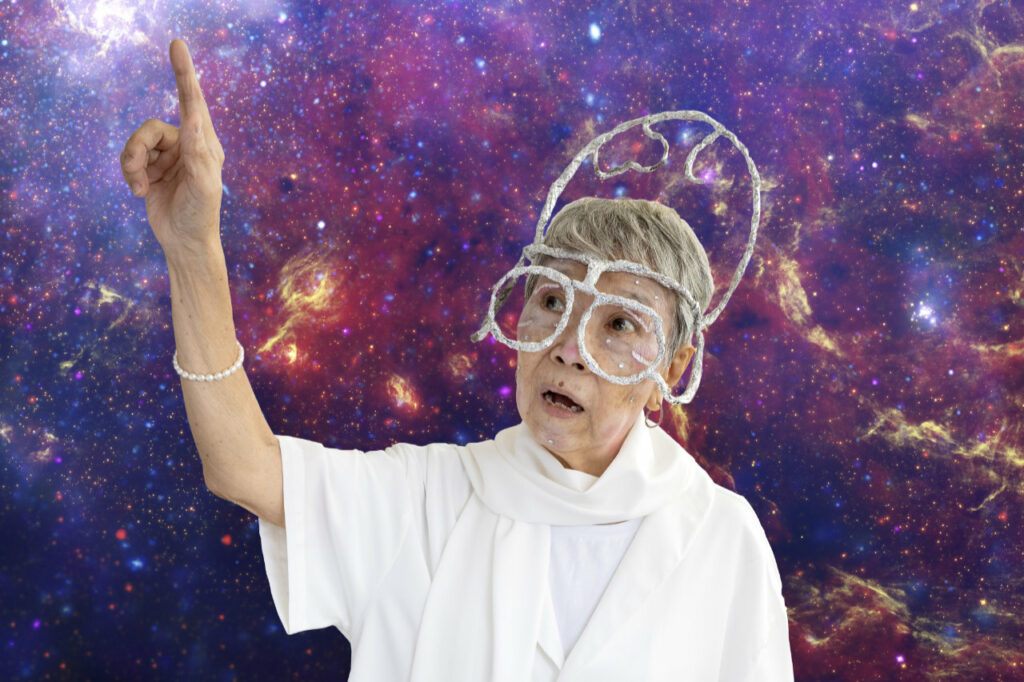 Senior collaborator, Lim Soh Joo, in a chrome coloured headpiece against a galaxy backdrop with her right hand raised and right index finger pointing upward.