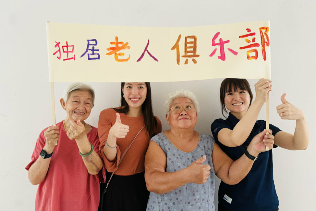 Senior collaborators, Yim Fong and Tong Cheng holding up a banner with 2 Yishun Health staff with text in Chinese: Single Seniors’ Club