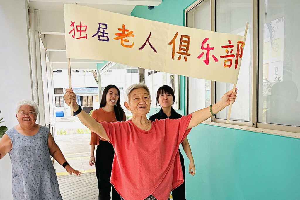 Senior collaborator, Tong Cheng, holding a banner with text in Chinese Single Seniors’ club. Senior collaborator, Yim Fong and 2 Yishun Health staff walking behind.