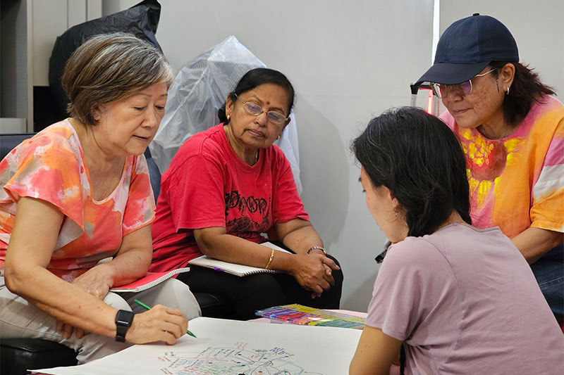 A senior participant writes on a large piece of paper with a colour pen as 2 other senior participants look on with an artist facilitator around a table
