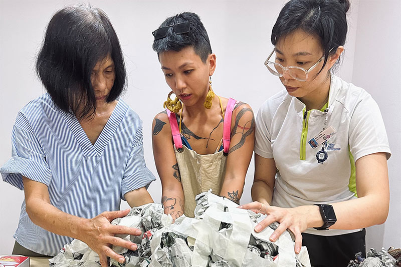 A senior participant, an artist facilitator and a healthcare worker are standing side by side with their hands on a paper mache sculpture